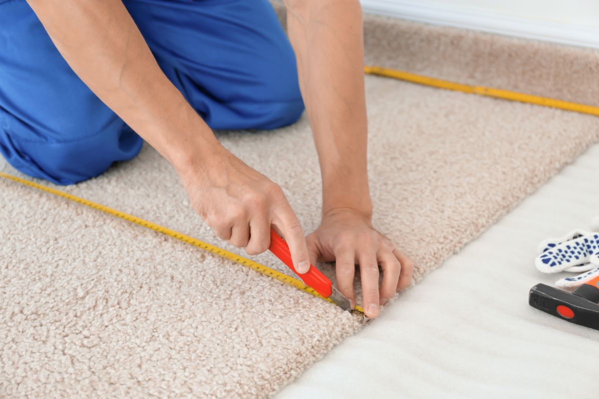 A person's hands are measuring and cutting carpet with a utility knife, surrounded by flooring tools on soft, light-colored carpet.
