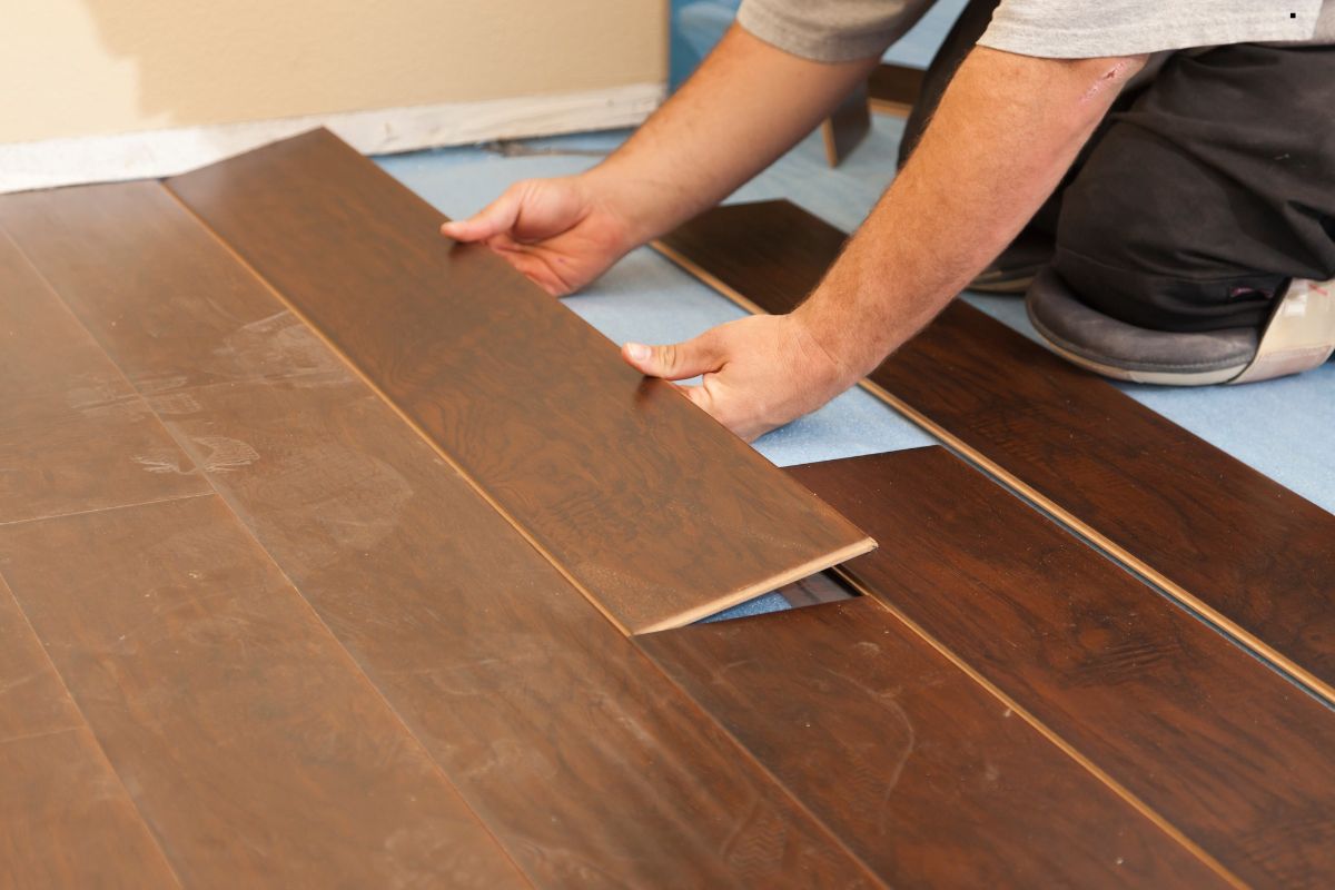 A person installs dark wooden flooring, carefully placing a new plank into position on a protective mat.