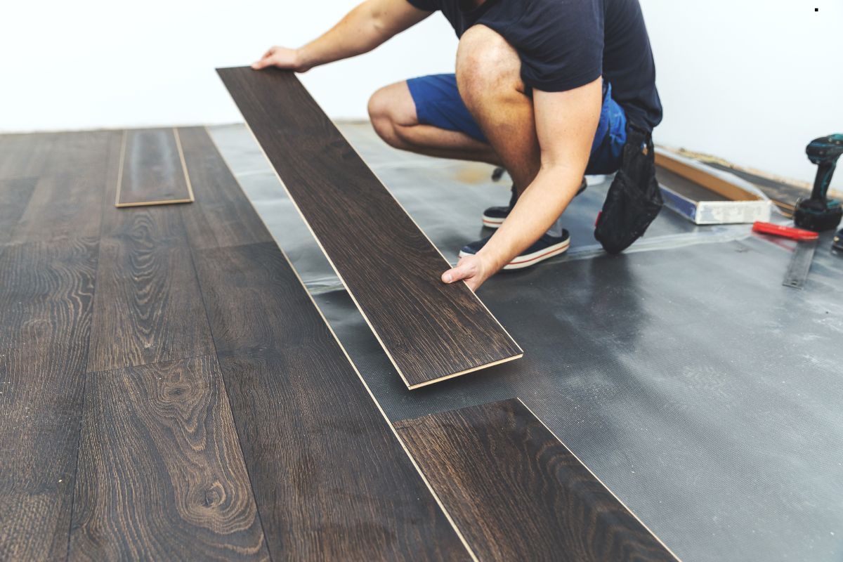 A person wearing a navy shirt and shorts is laying dark wooden floor planks in a room under renovation. Tools are in the background.