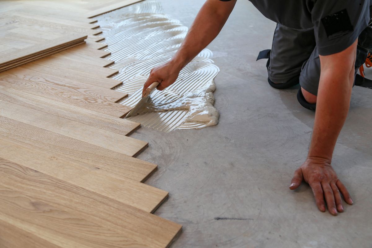 A person kneels on the floor, applying adhesive to wood planks for installation. The smooth adhesive creates wave patterns.
